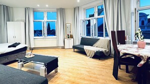 A modern hotel room with a bed, sofa, dining table, and large windows offering a city view. - Cozy 4BR Apartment Near Kaufpark (Vienna)