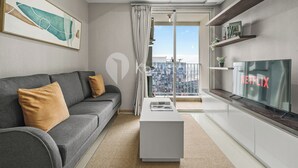 A modern living room with a grey sofa, a coffee table, a large window with a view of a cityscape, and a flat-screen TV mounted on a wooden shelf. - Nahara by Kozystay - Kuningan (Jakarta)
