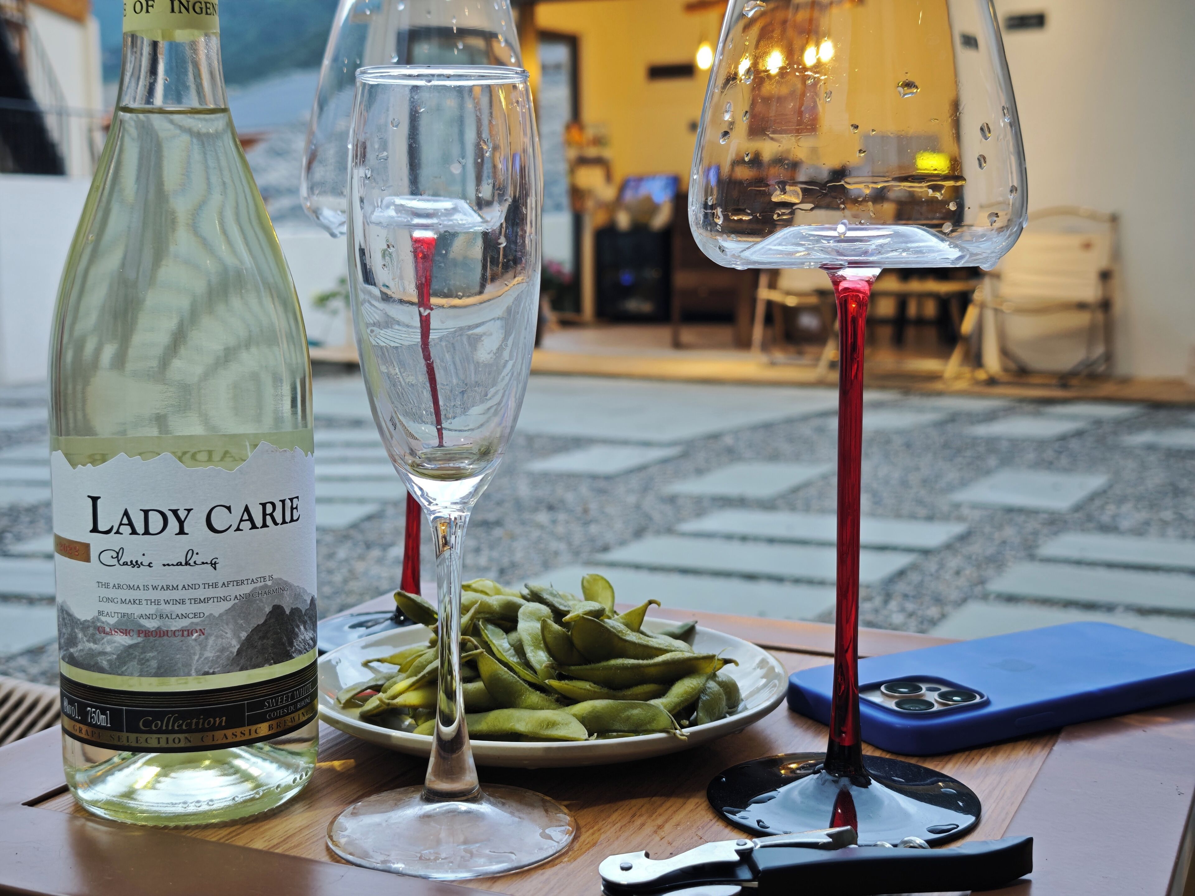 A bottle of Lady Carie wine, two glasses with red wine, a plate of green beans, and a smartphone on a wooden table.