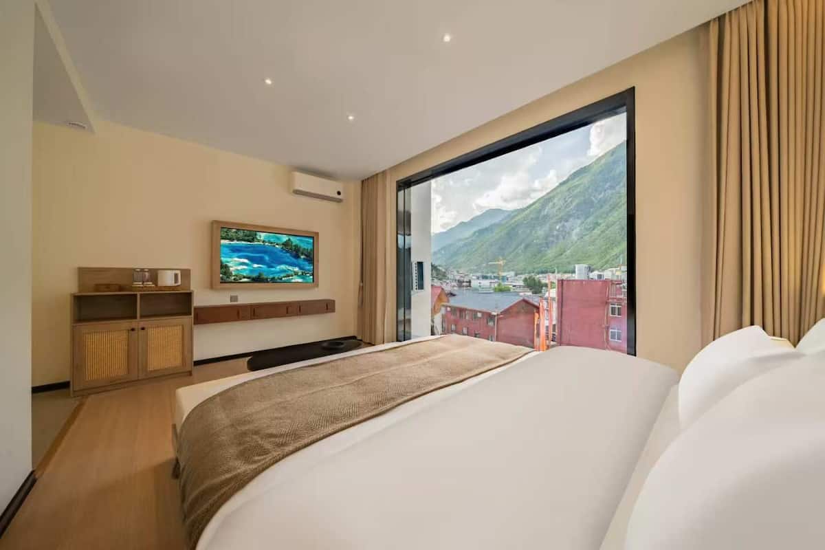 Ein Hotelzimmer mit einem großen Bett, einem Fernseher an der Wand und Blick auf Berge und Gebäude.