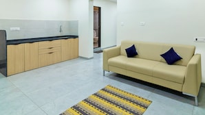 A modern living room with a beige sofa and a patterned rug, a kitchen with wooden cabinets and a black countertop, and a doorway leading to a bathroom. - Tribe Moro Balewadi  (Paud)