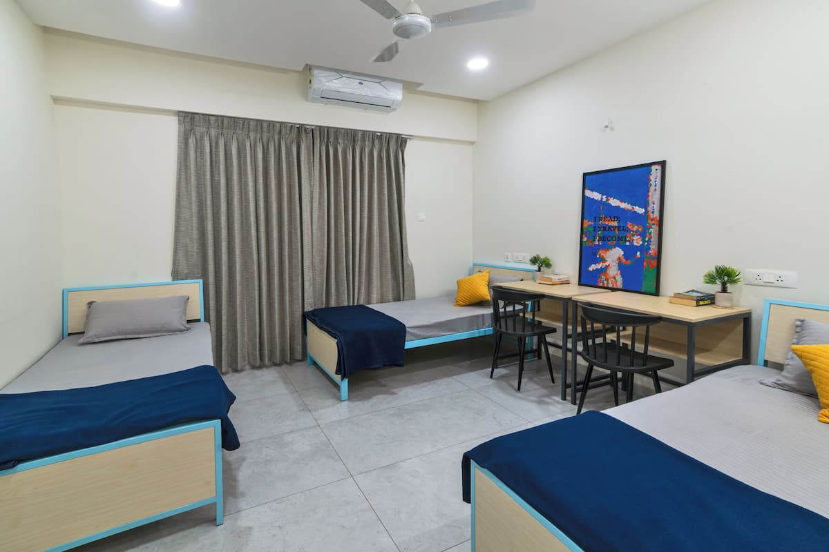 A dormitory room with two single beds, a desk with a chair, a ceiling fan, an air conditioning unit, and a poster on the wall.