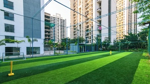 A well-maintained sports court with a net, surrounded by greenery and tall buildings. - Tribe Moro Balewadi  (Paud)