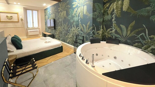 A hotel room with a large bathtub, a bed with a green headboard, a wooden bench, and a tropical wallpaper design. - Atlas Apartments  (Bergamo)