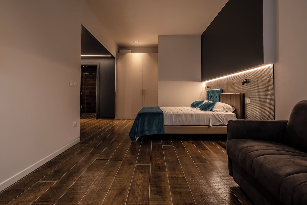 A modern bedroom with a bed, a dark sofa, and a wooden floor.