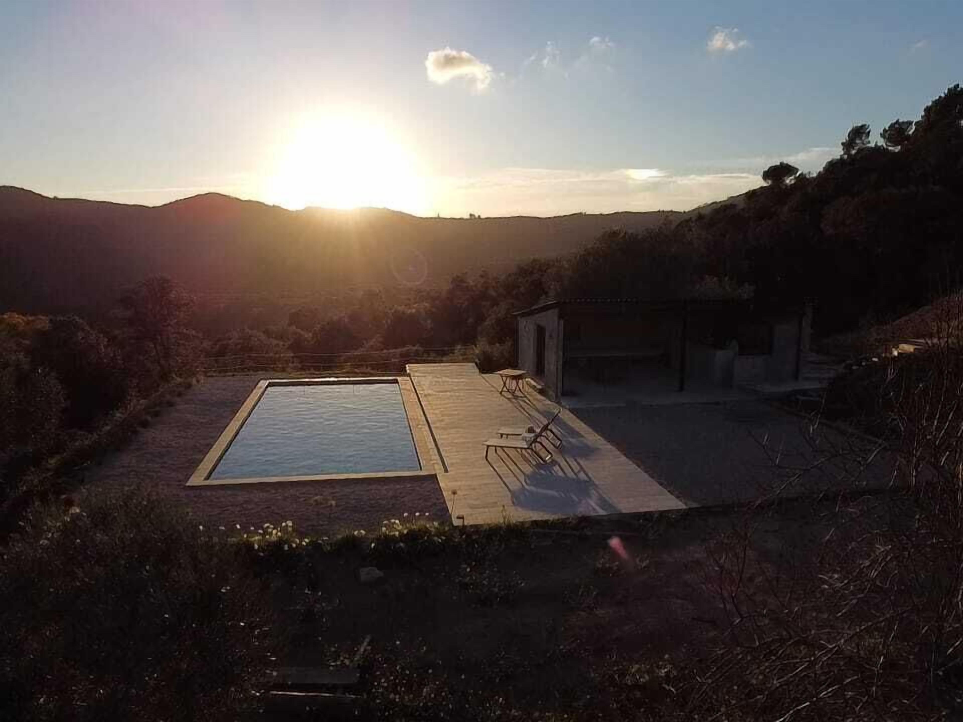 Piscina retangular com um deck de madeira e espreguiçadeiras, cercada por colinas e um céu limpo.