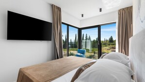 Ein modernes Schlafzimmer mit einem großen Bett, einem flachbildfernseher an der Wand und einer Glasschiebetür, die auf einen Balkon mit Blick auf Bäume und einen klaren Himmel führt.