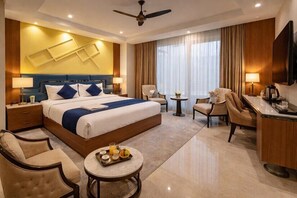 Ein Hotelzimmer mit einem großen Bett, zwei Sesseln, einem Schreibtisch, einem Fernseher und einem Deckenventilator.