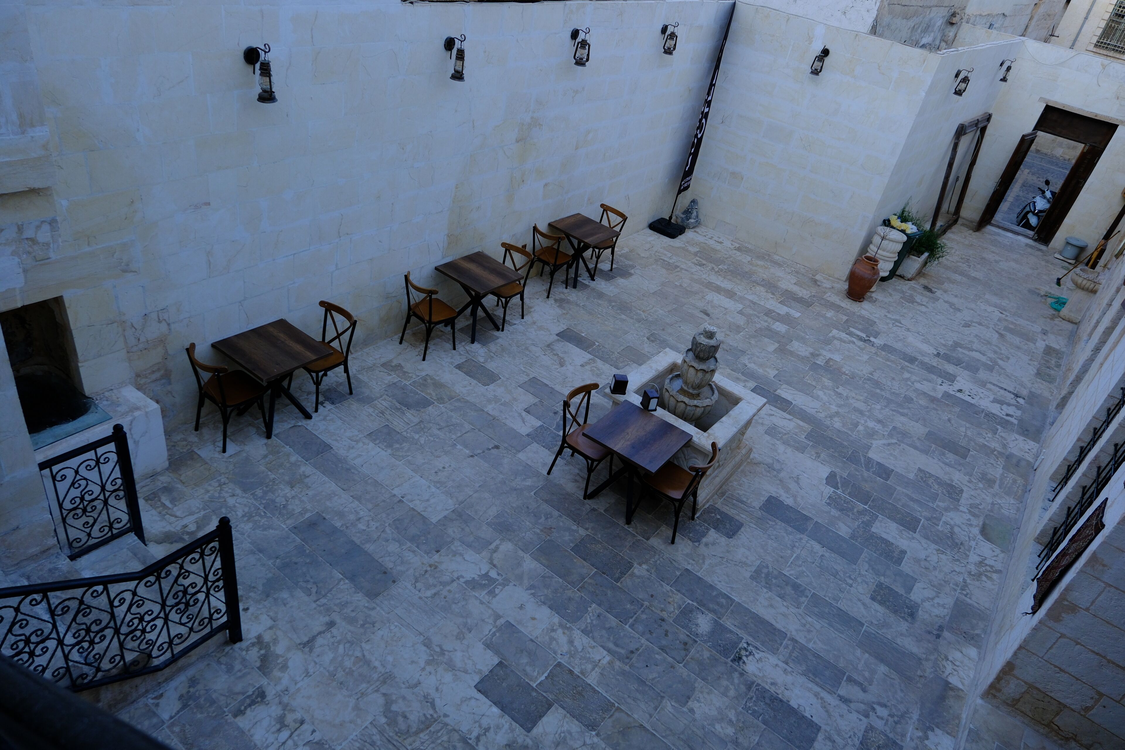 A courtyard with stone flooring, wooden tables and chairs, and lanterns on the walls.
