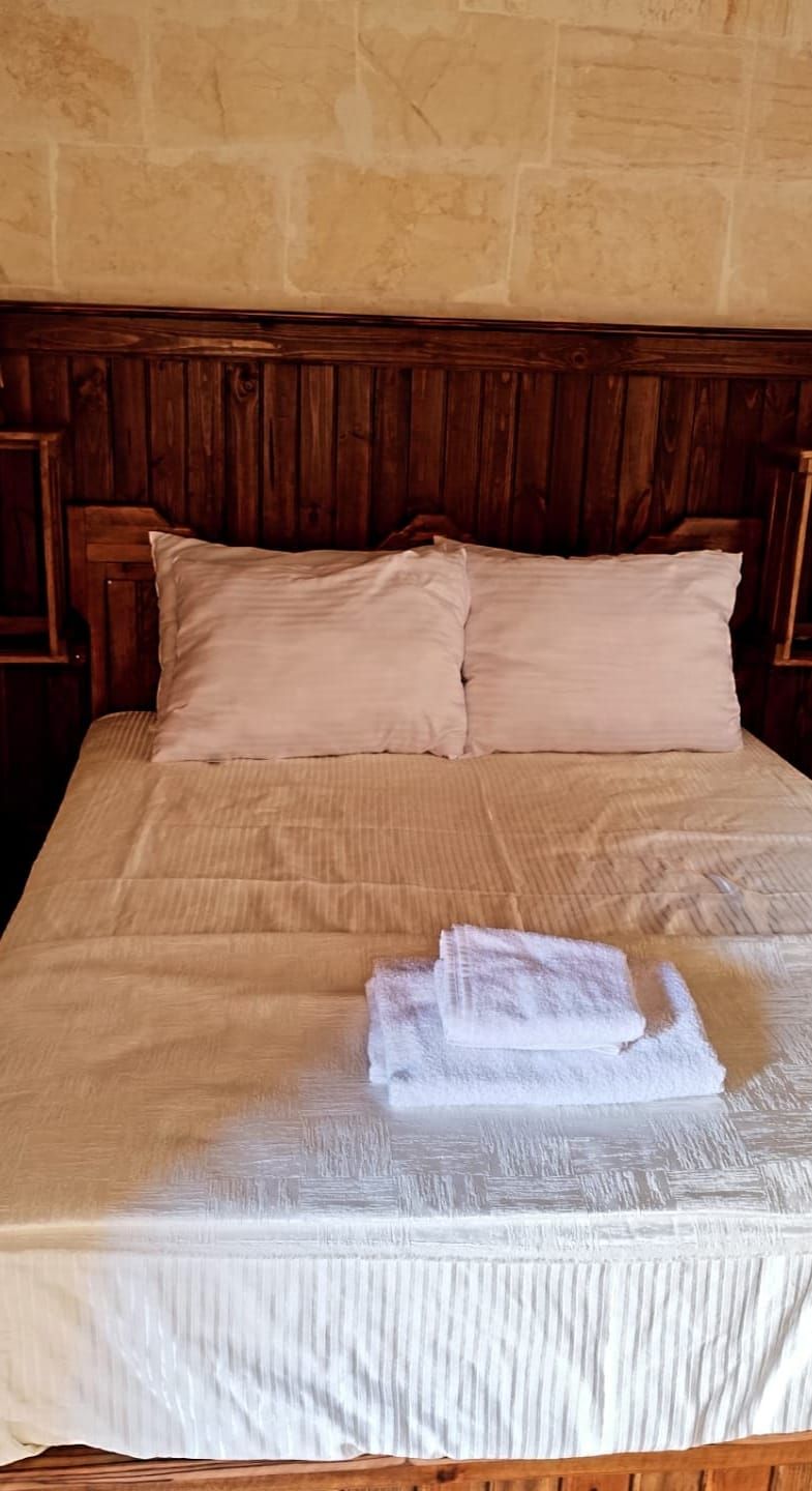 Un lit bien fait, avec une tête de lit en bois, une serviette blanche pliée posée sur le lit, et un mur de pierre en arrière-plan.