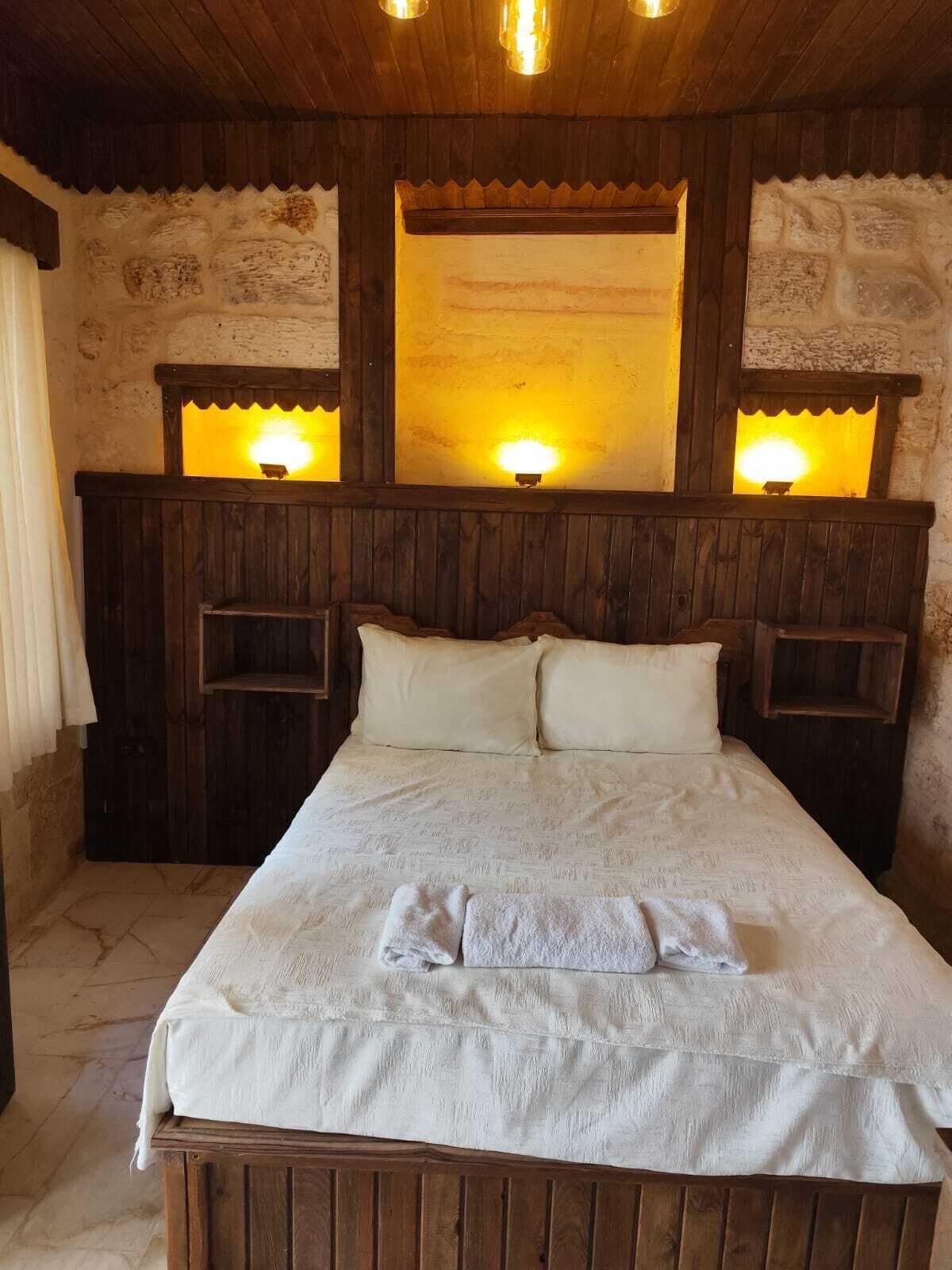 Una cama de madera con una colcha blanca y dos almohadas, flanqueada por mesitas de noche de madera con lámparas de pared.
