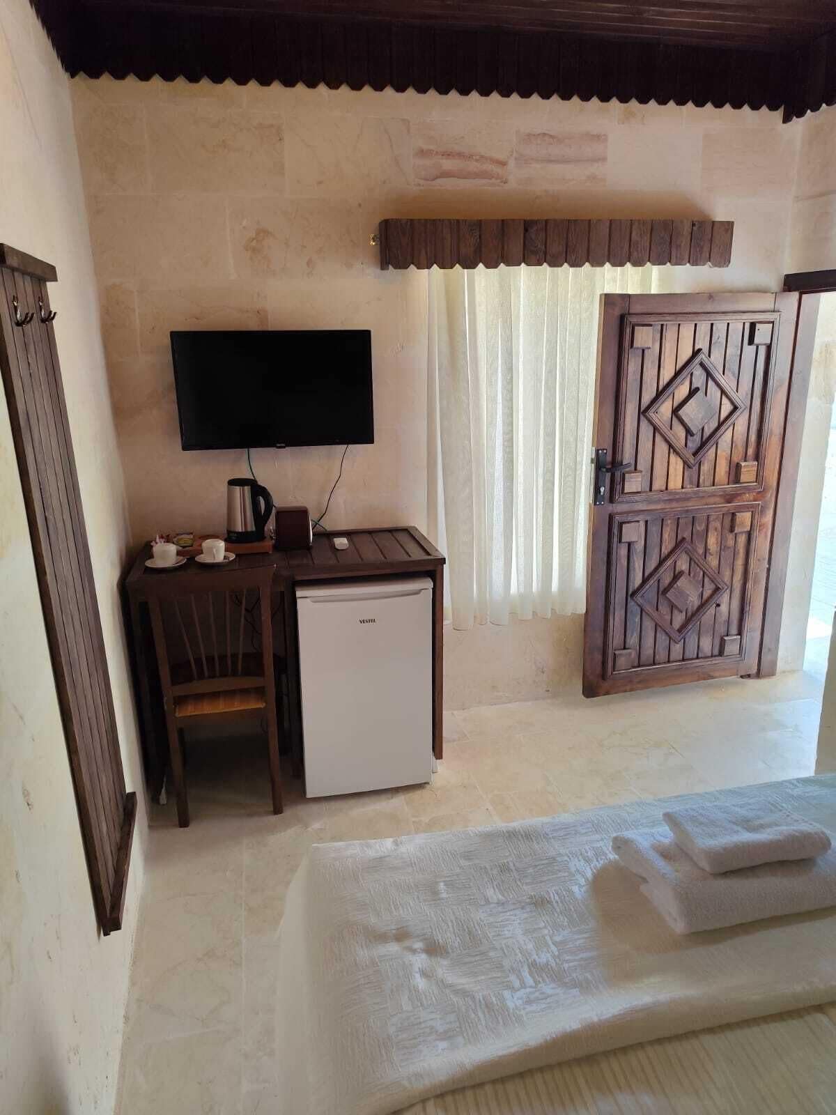 Quarto com televisão, pequeno frigorífico, secretária de madeira, cadeira, cama com edredão branco e porta de madeira decorativa.