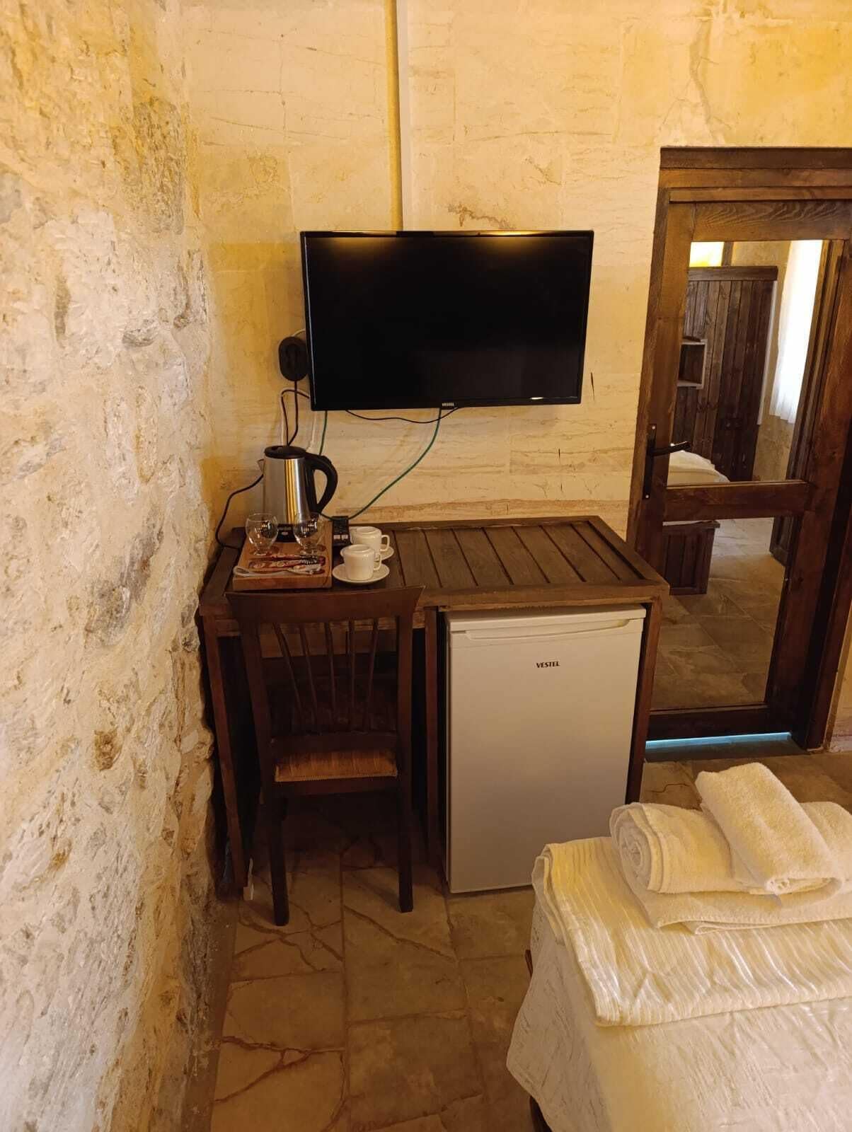 A small room with a wooden table, a television mounted on the wall, a small refrigerator, and a bed with a white blanket.