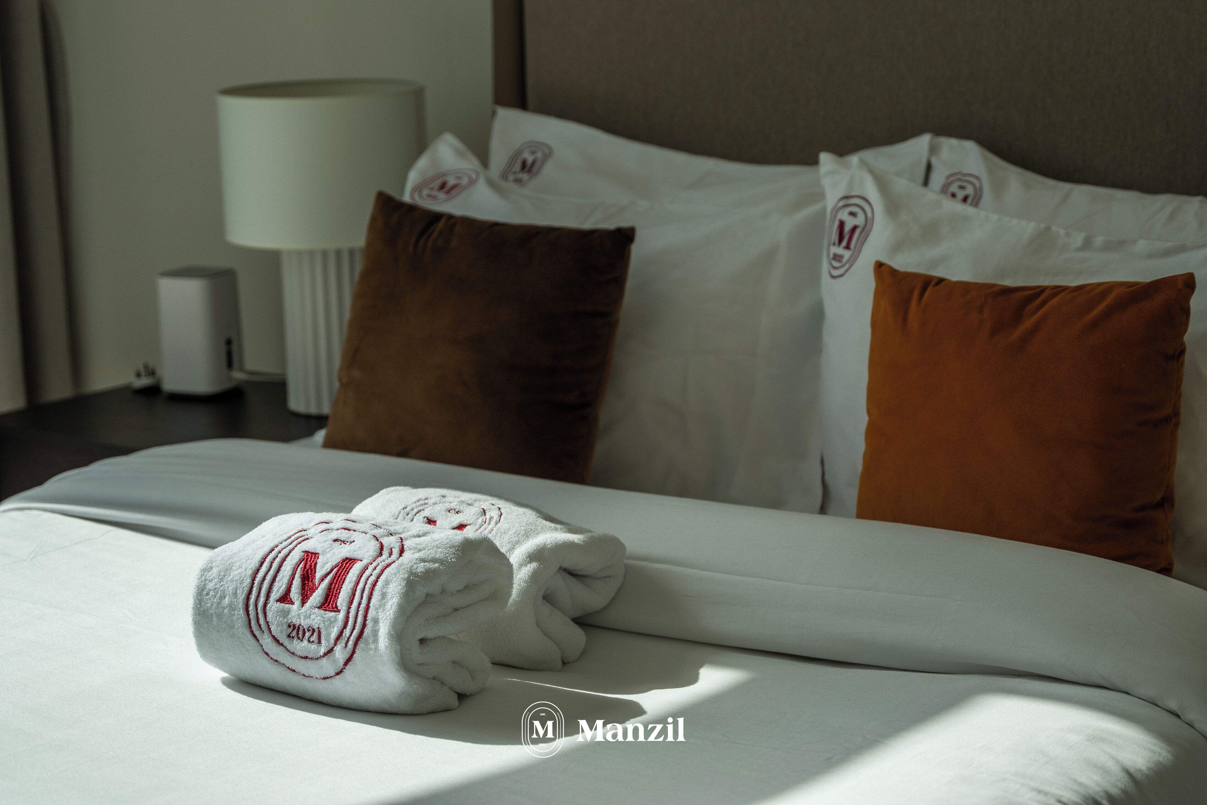 A neatly made bed with a white duvet, brown and white pillows, and a folded towel with a logo.