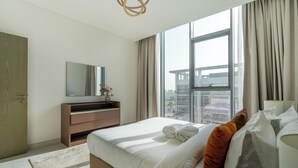 A modern hotel room with a large bed, a wooden dresser, a mirror, and a view of a cityscape through large windows. - Manzil - Lux 1BR minutes to Dubai Mall (Dubai)