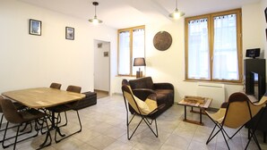 A spacious living area with a wooden dining table, two sofas, a coffee table, and a TV. There are two windows with curtains, a clock on the wall, and a lamp. - Beautiful F3 with parking and pets allowed, near thermal baths (Mont-Dore)