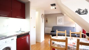 Ett kompakt kök med tvättmaskin, ett matbord med stolar, en grå soffa och en vägg dekorerad med en bild av en hjort och ett träd.