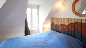 Ett sovrum med ett sluttande tak, en sänggavel i trä med ett bergsmotiv, en rund spegel och ett fönster med gardiner.