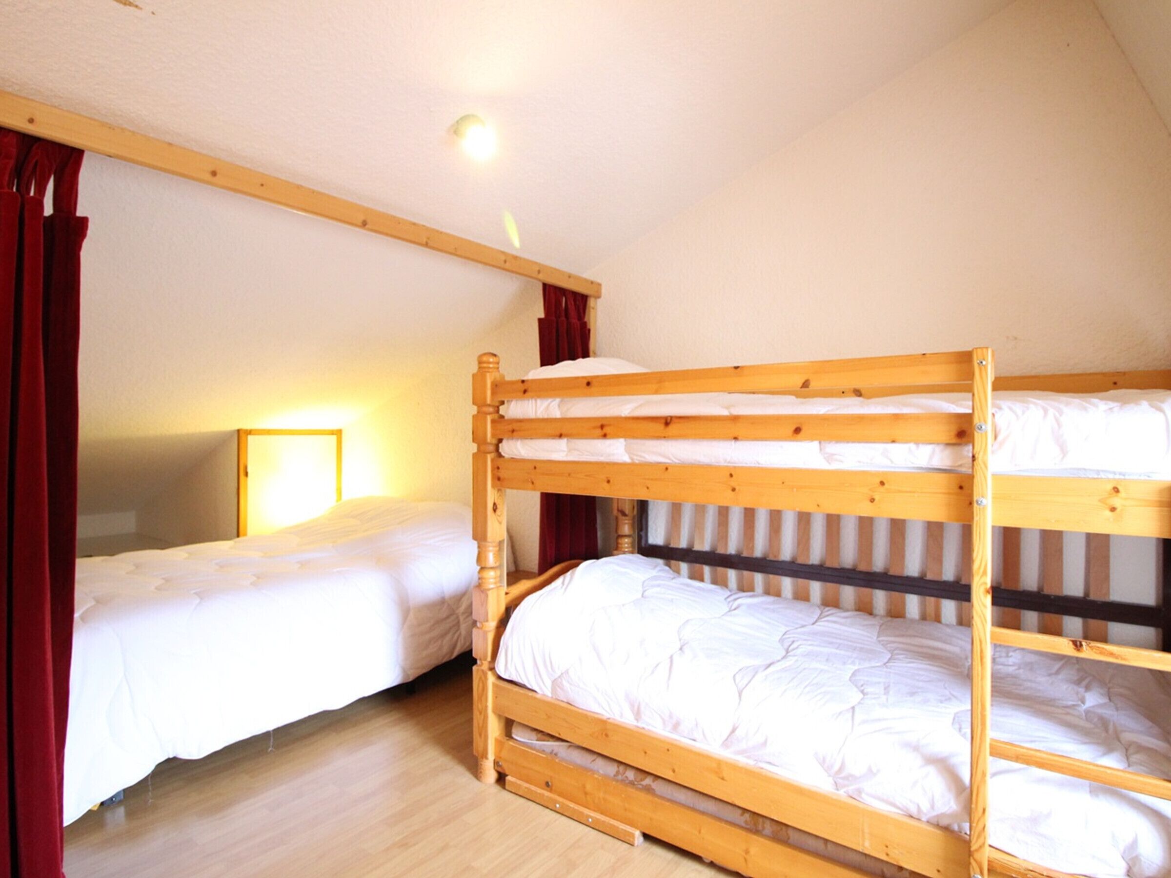 A room with two bunk beds, wooden flooring, a mirror on the wall, and a window with curtains.