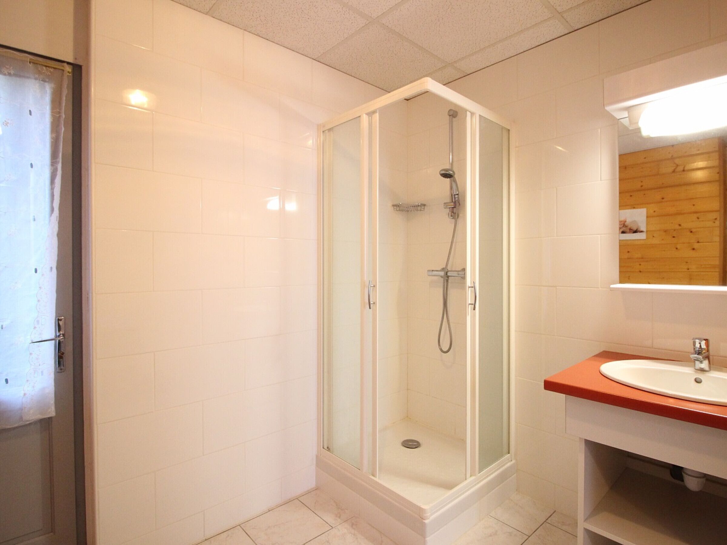 Ein modernes Badezimmer mit einer gläsernen Duschkabine, einem weißen Waschbecken mit orangefarbener Arbeitsplatte und einer Wandverkleidung aus Holz.