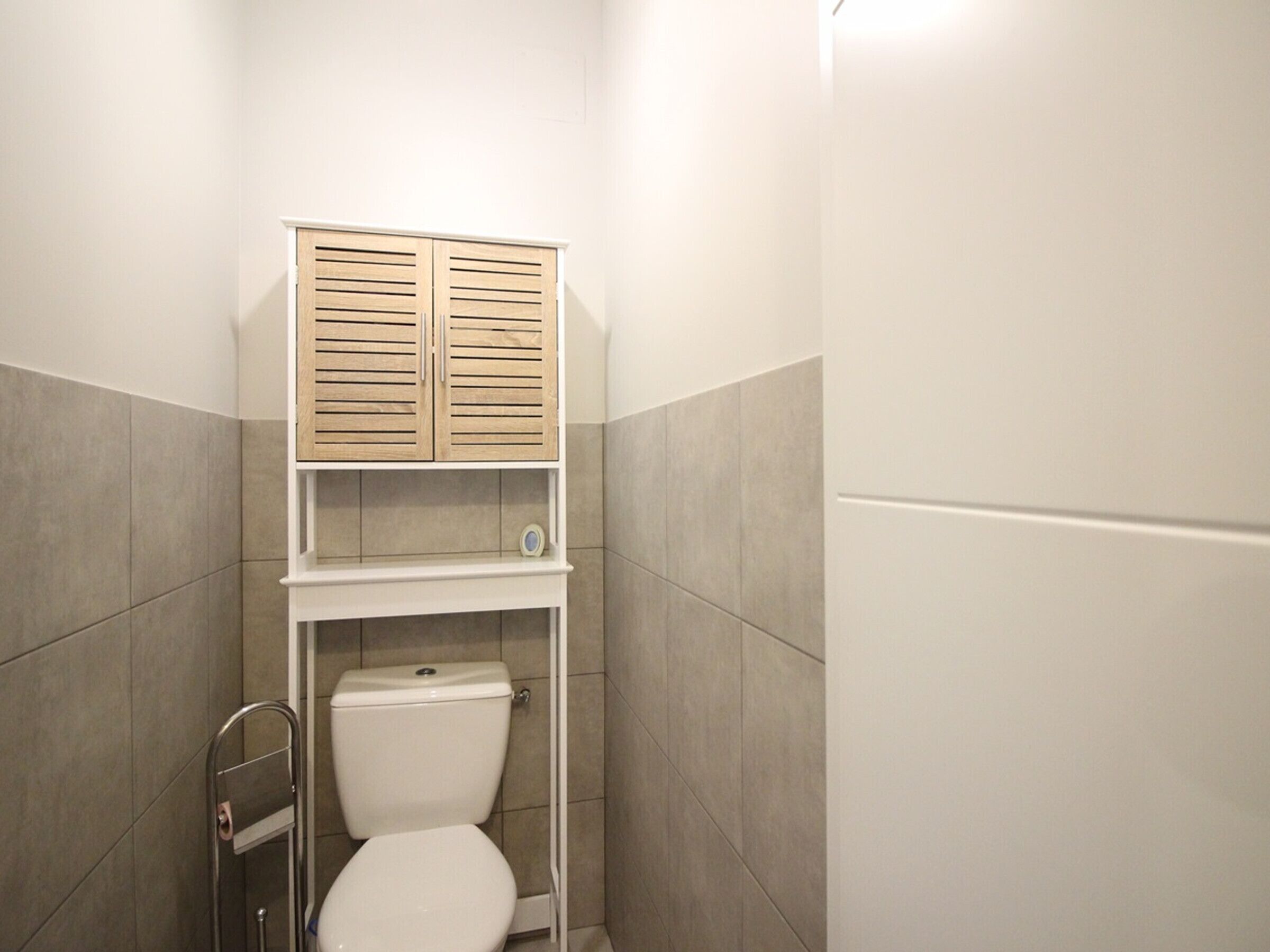 Une petite salle de bain fermée, comprenant des toilettes, une étagère avec une horloge et un meuble en bois placé au-dessus des toilettes.