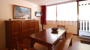 Uma sala de jantar com uma mesa de madeira, cadeiras, um banco, uma televisão sobre um suporte, um quadro na parede e uma janela com cortinas.