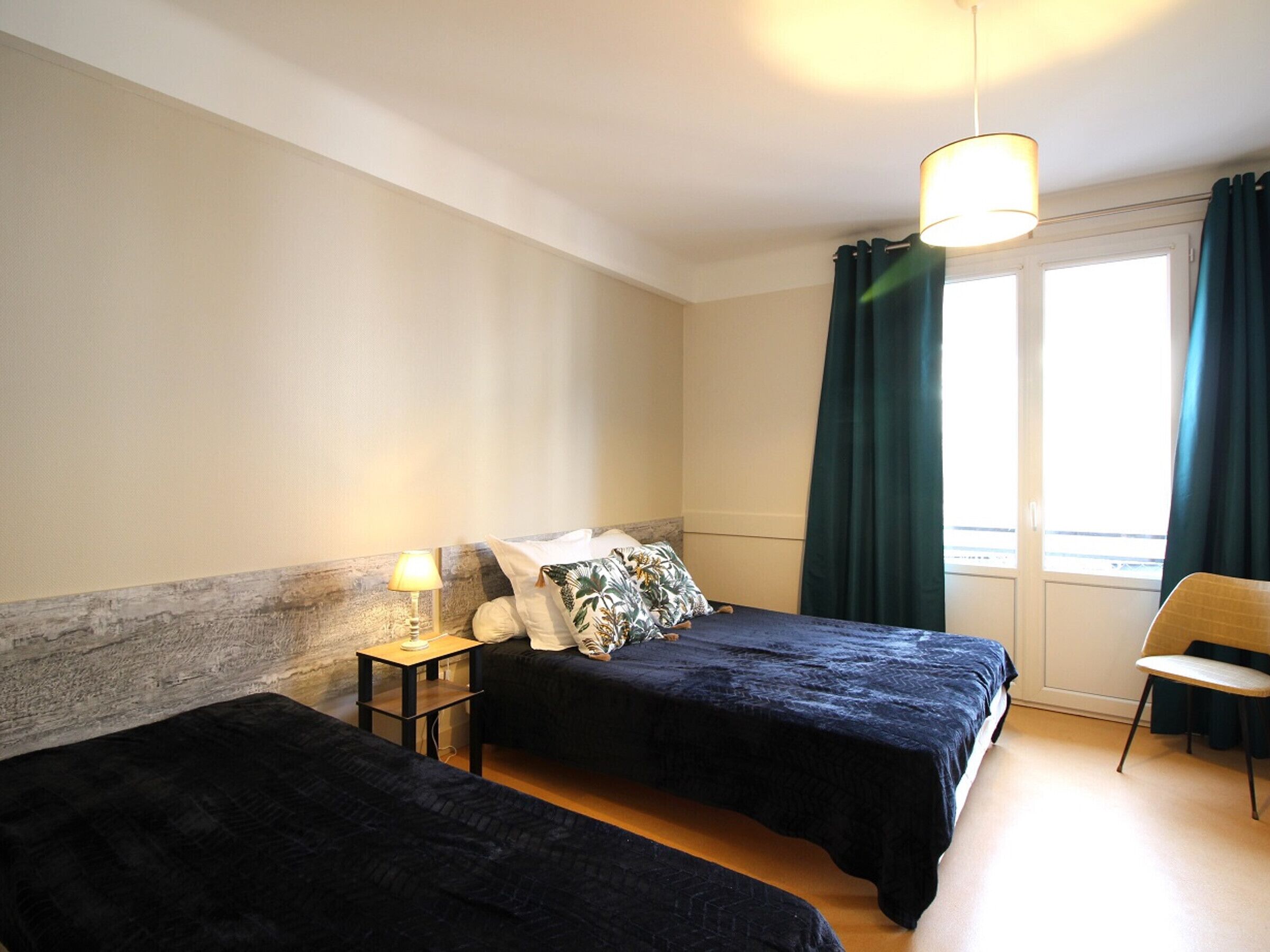 A hotel room with two beds, a nightstand with a lamp, a chair, and a window with curtains.