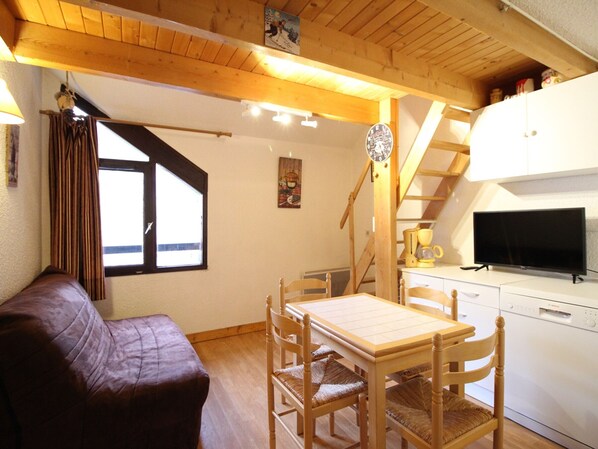 Une pièce chaleureuse avec un plafond en bois, un canapé, une table à manger avec des chaises, un coin cuisine équipé d’un lave-vaisselle et une fenêtre avec des rideaux.