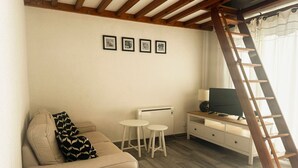 Una habitación con un sofá, una pequeña mesa, un televisor sobre un soporte de madera, una escalera y un radiador.