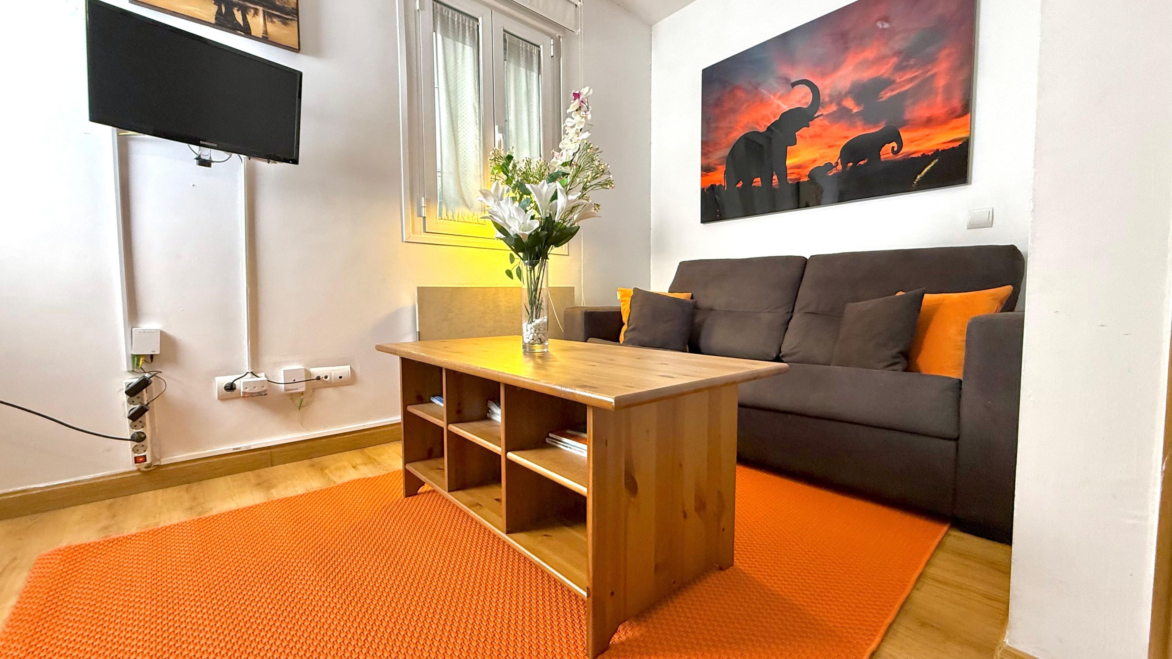 Un salón con un sofá, una mesa de centro de madera, un florero con flores, un cuadro en la pared y un televisor montado en la pared.