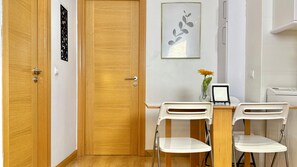 A room with a wooden door, a white desk with two chairs, a vase with a flower, and a framed picture on the wall. - Alaia Gran Via Seasonal Living (Madrid)
