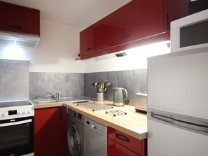 Eine kleine Küche mit roten Schränken, einer Edelstahl-Spüle, einer Waschmaschine und einem Kühlschrank.