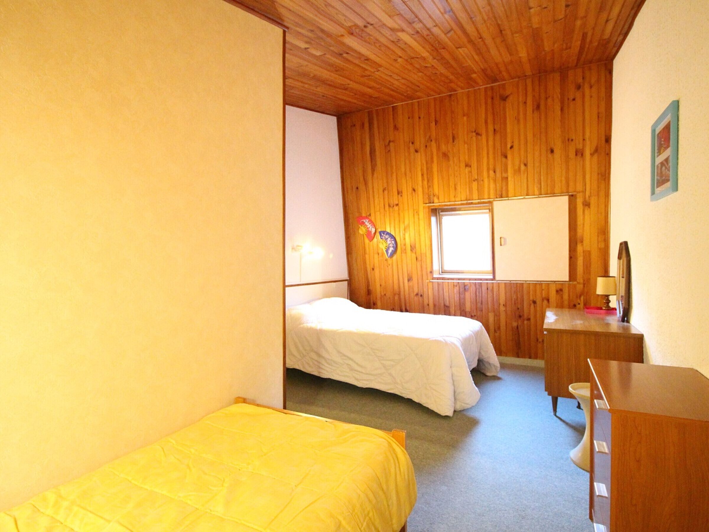 A room with two beds, a wooden ceiling, a window, and a desk with a mirror.