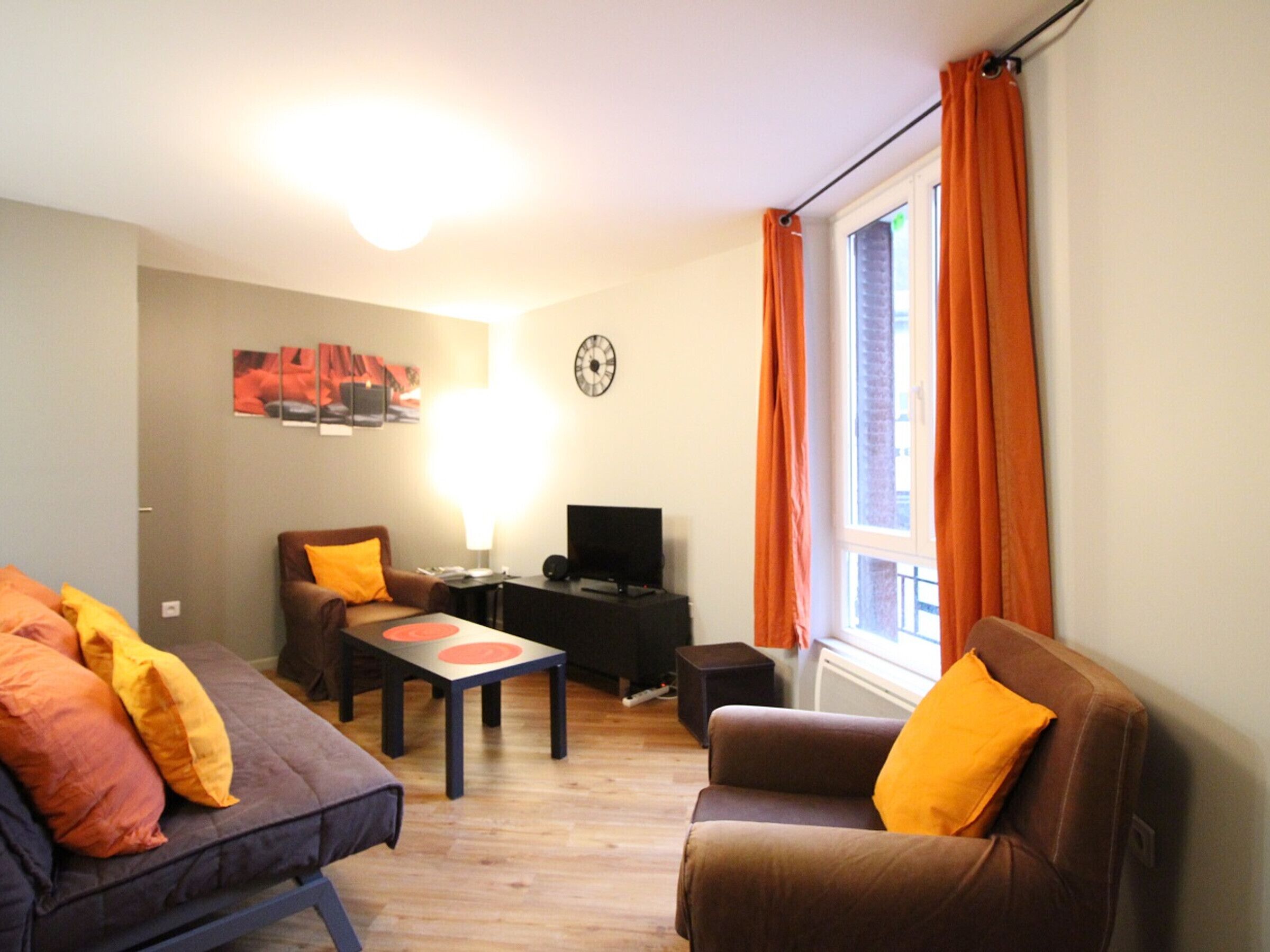 Ein Wohnzimmer mit einer Couch, einem Sessel und einem kleinen Tisch mit Lampe. Ein Fernseher steht auf einem Schrank und eine Uhr hängt an der Wand.