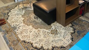 Una sala de estar moderna con un sofá de cuero negro, una mesita blanca y una mesa de centro de madera. Hay una alfombra grande con un diseño llamativo en el suelo, y la habitación tiene un diseño minimalista con un estante de madera y un espejo en la pared.