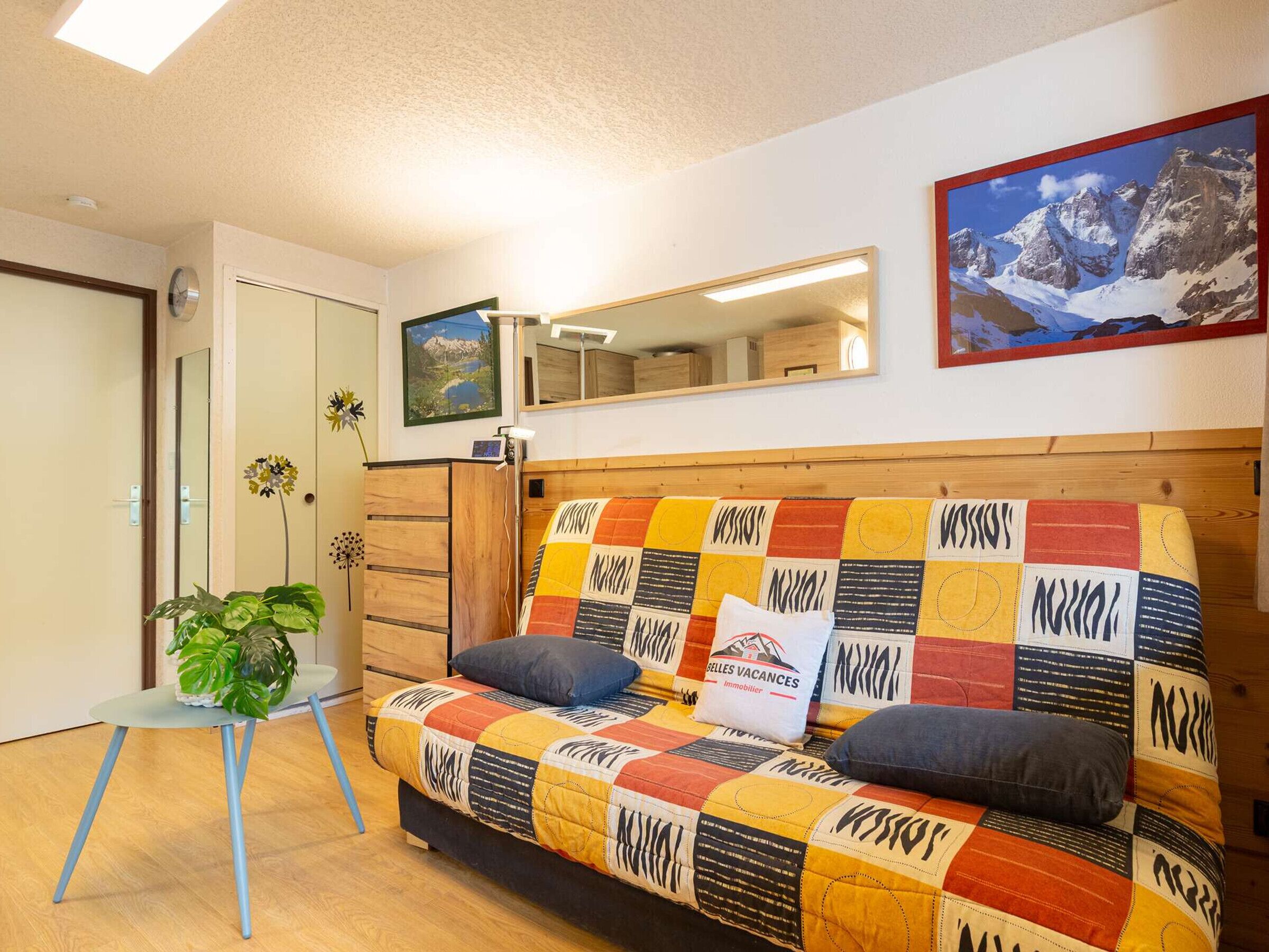 A cozy room with a colorful patchwork sofa, a small table with a plant, a mirror, and a framed picture on the wall.