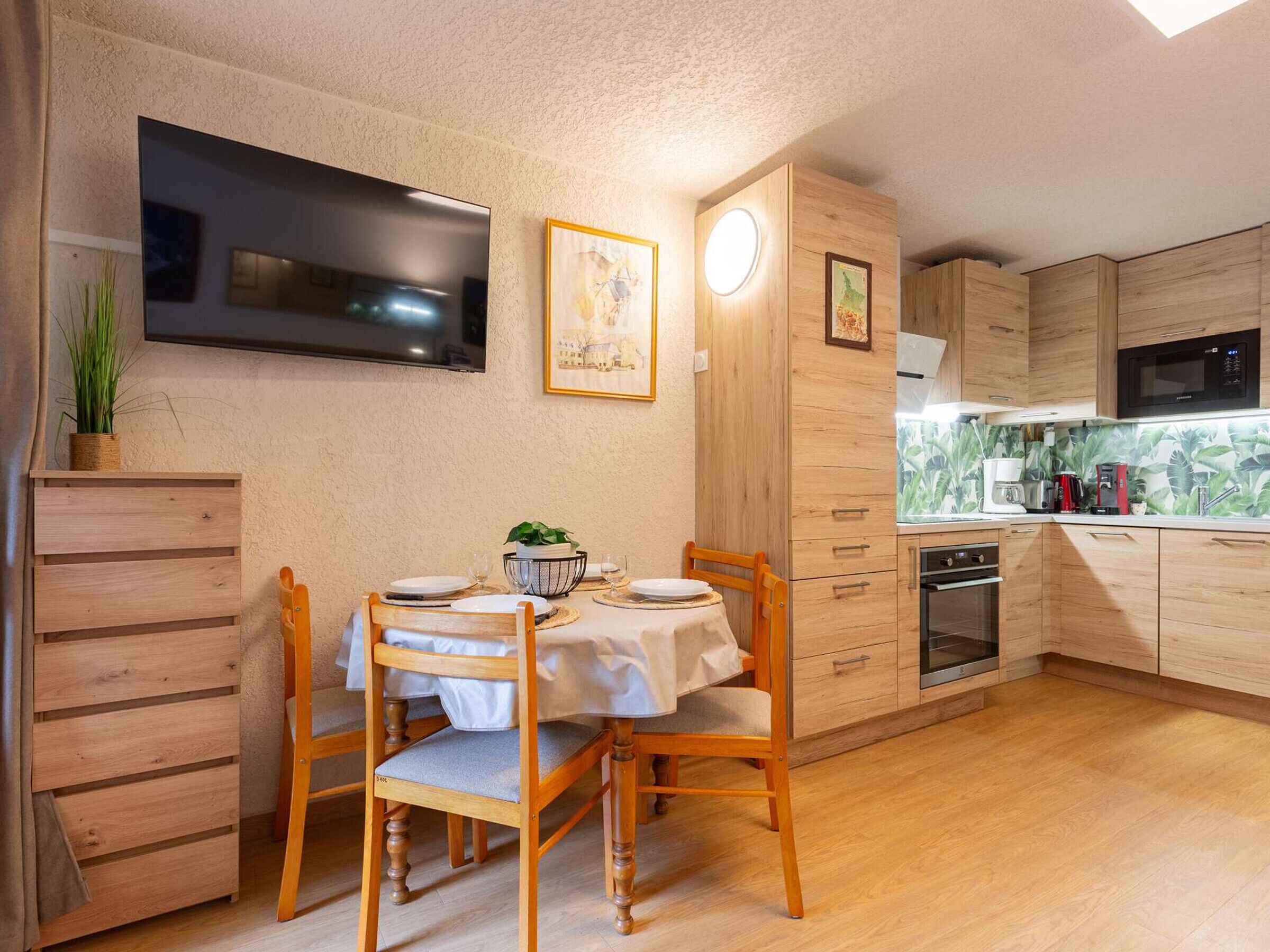 A compact kitchen with wooden cabinets, a dining table with chairs, a built-in oven, and a flat-screen TV mounted on the wall.