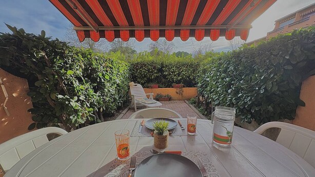 An outdoor dining area with a table set for two, surrounded by lush greenery and a red-striped awning.