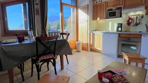 Zona pranzo con tavolo e sedie, cucina completa di elettrodomestici e vista sulle montagne attraverso ampie finestre.