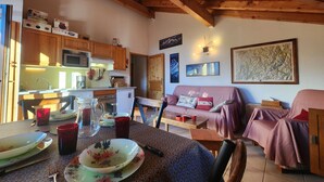 Una cucina accogliente con travi in legno, un tavolo da pranzo apparecchiato con piatti e tazze, e una zona soggiorno con divano e cuscini.