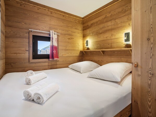 Quarto com painéis de madeira, uma cama, duas almofadas e uma janela com cortina.