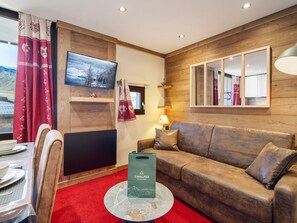 A cozy room with a wooden wall, a television mounted above a flat-screen TV, a sofa, a dining table with chairs, and a red carpet. - Ski-in/Ski-out Apartment in Val Thorens with WIFI (Val Thorens)