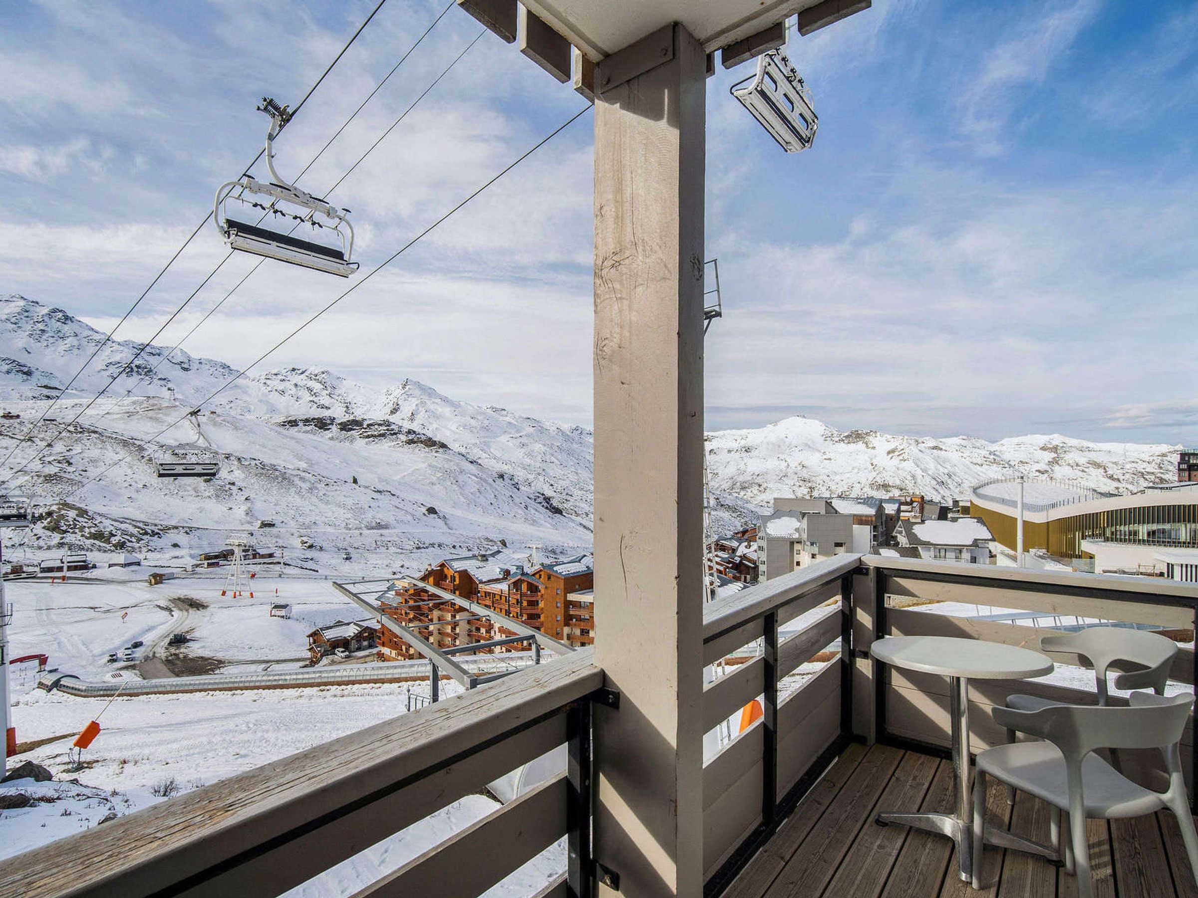 A balcony with a view of a ski lift, snow-covered mountains, and a building with a modern design.
