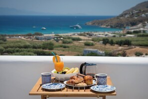 Ein Frühstücksbuffet mit Croissants, Obst und Kaffee auf einem Holztisch, im Hintergrund ein schöner Blick auf Meer und Hügel.
