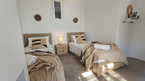 A bedroom with two beds, a bedside table with a lamp, and a small shelf with decorative items. - Mykonos4Islands Villa "Athena" with shared pool - 5 min walking from the beach (MYKONOS)