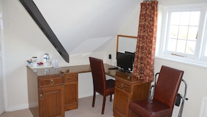 Ein Zimmer mit Schreibtisch, zwei Stühlen, einer kleinen Küchenzeile, einem Fenster mit Vorhang und einer Heizung.