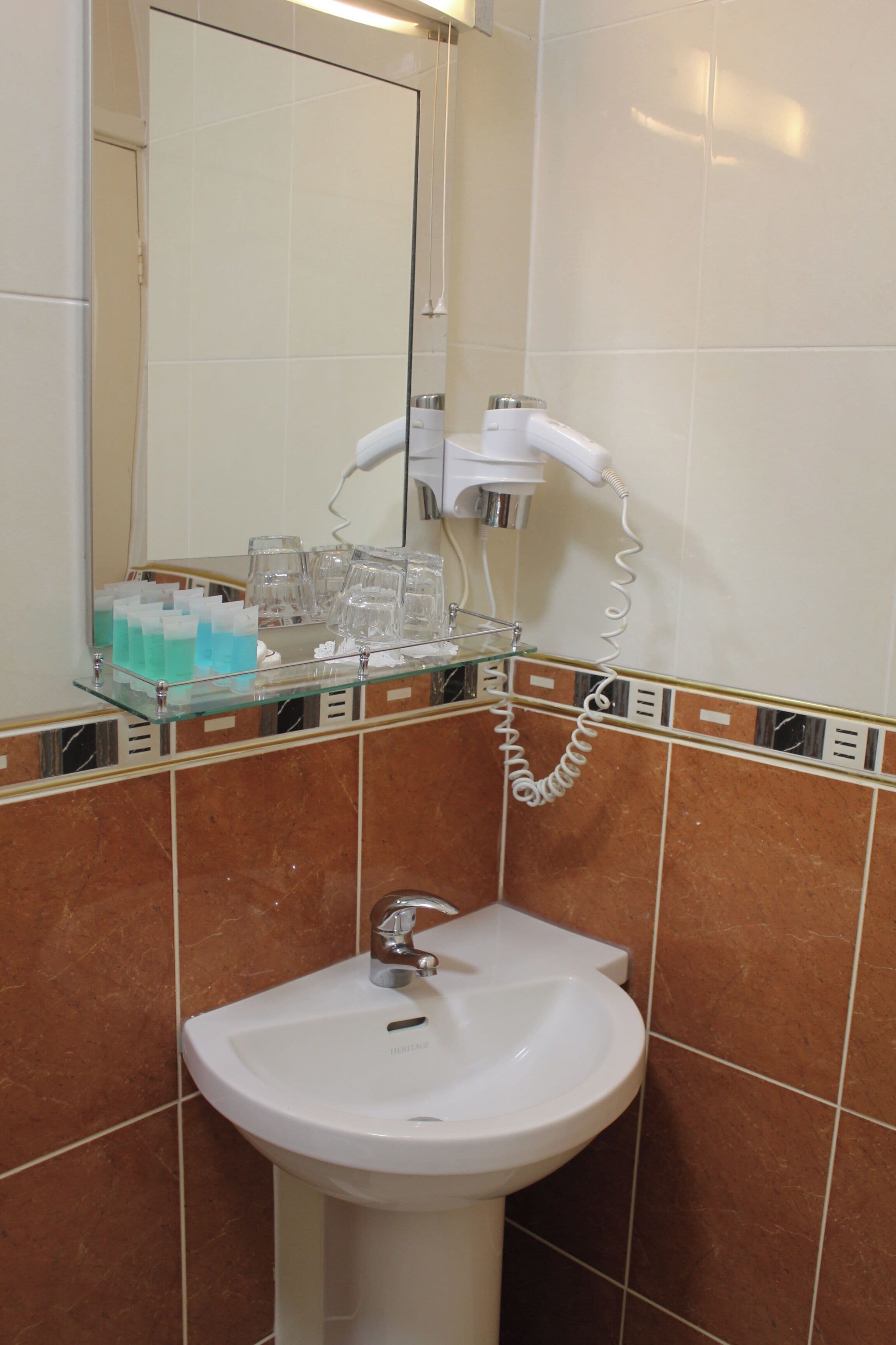 Une salle de bain avec un lavabo blanc, une étagère en verre où sont rangés les articles de toilette et un sèche-cheveux fixé au mur.