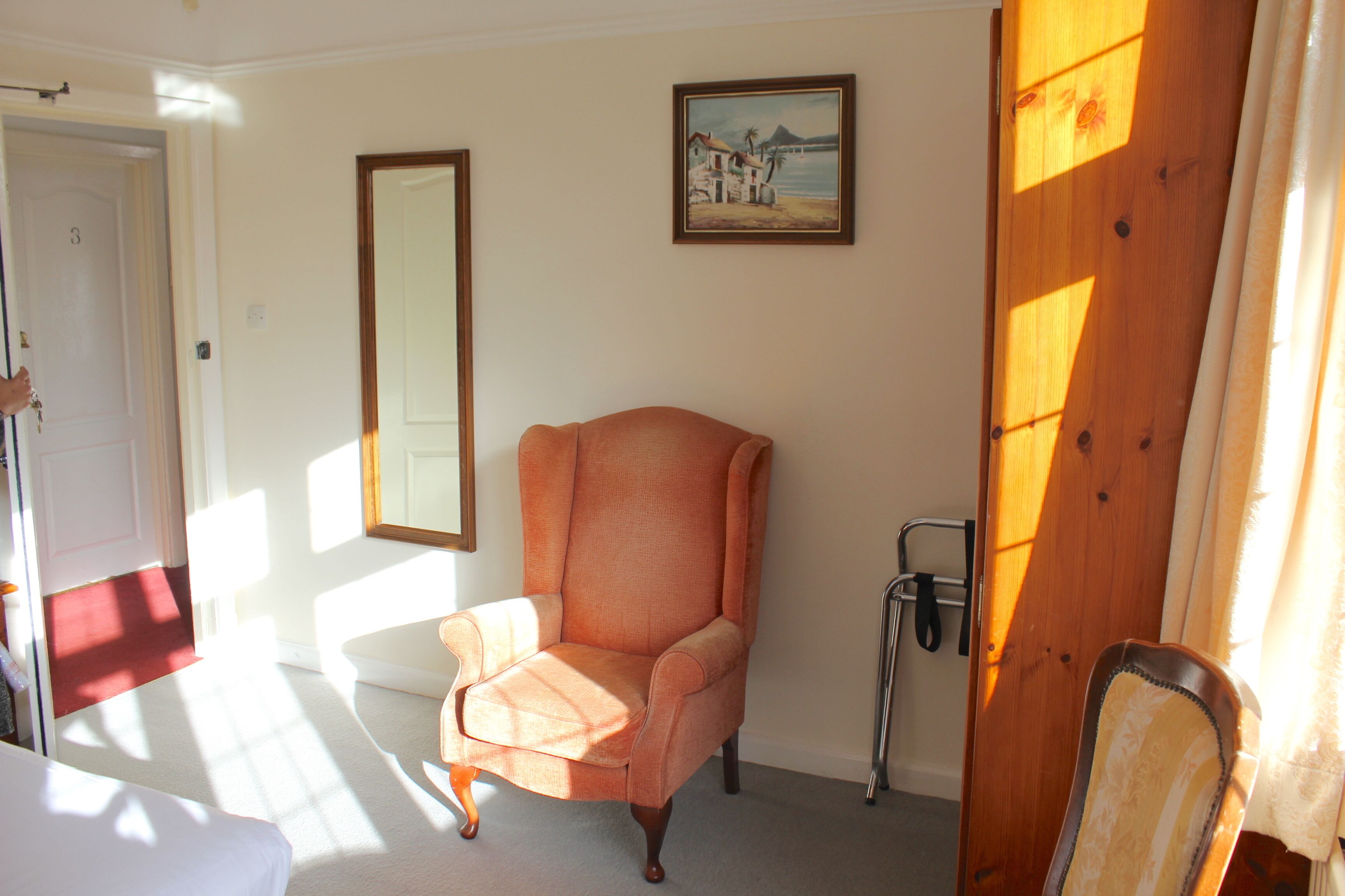 Una habitación con un sillón naranja, una puerta de madera, un cuadro enmarcado en la pared y una ventana por donde entra la luz del sol proyectando sombras.