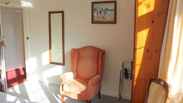 Una habitación con un sillón naranja, una puerta de madera, un cuadro enmarcado en la pared y una ventana por donde entra la luz del sol proyectando sombras.