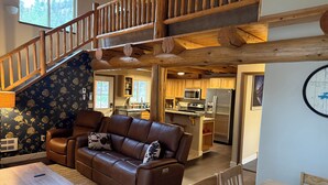 A spacious living area with a brown leather sofa, a wooden dining table, and a kitchen in the background. - Secluded 3 bed house in Kila with flowing creek, hot tub, with plenty of parking (Kila)